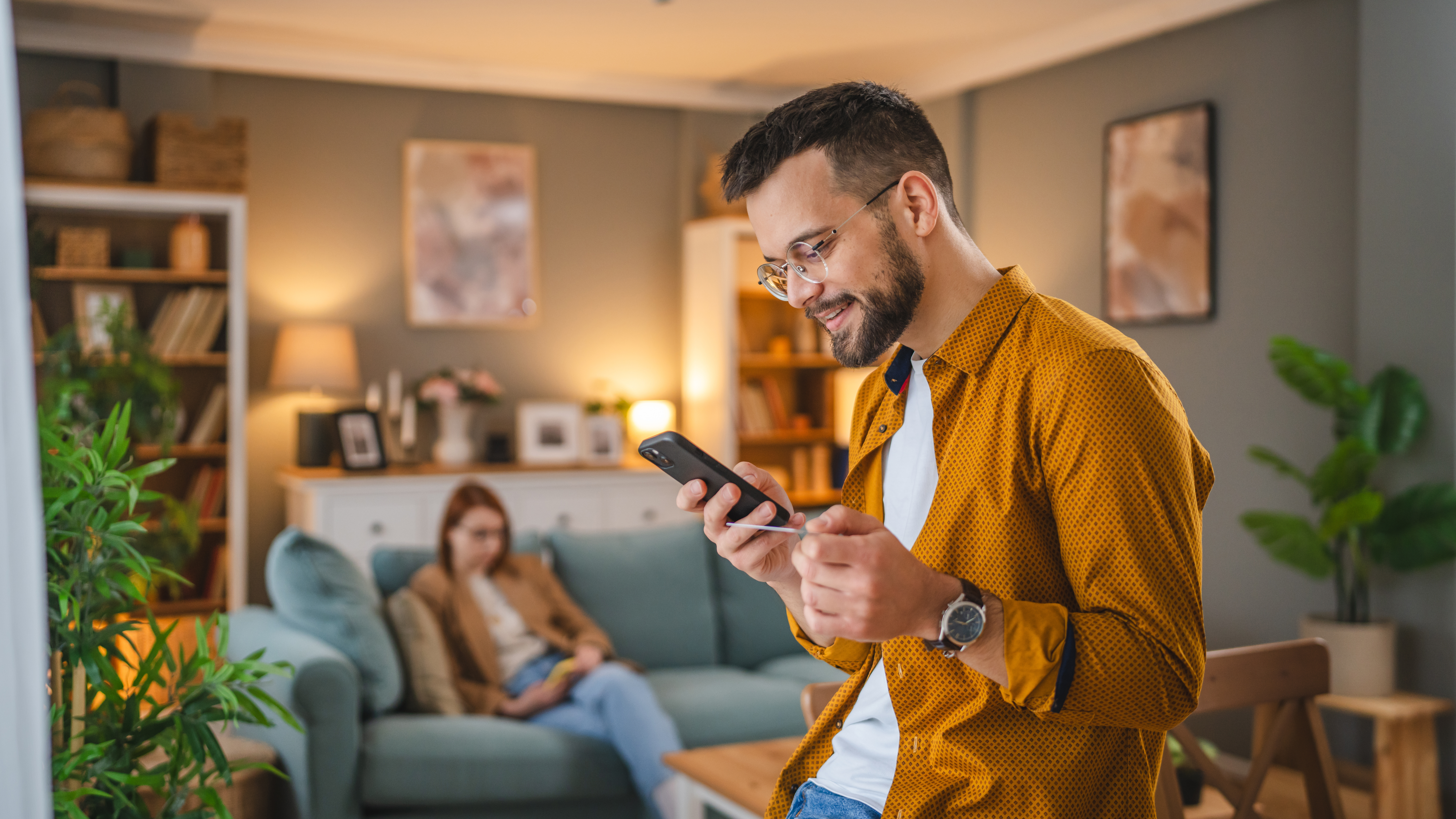 man doing online banking in his living room with wife in background. 